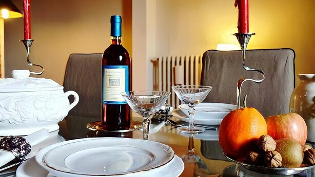 A dining table set with white plates, a bottle of wine, a white teapot, a martini glass, a fruit bowl with oranges and nuts, and two red candles.
