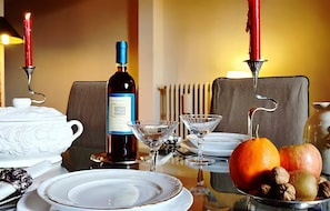 Ein Esstisch gedeckt mit weißen Tellern, einer Weinflasche, einer weißen Teekanne, einem Martini-Glas, einer Obstschale mit Orangen und Nüssen sowie zwei roten Kerzen.