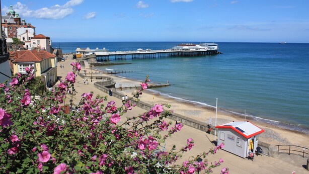 Eine Küstenstadt mit einem Pier, der ins Meer hinausführt, einer Promenade mit pinkfarbenen Blumen und einem kleinen weißen Gebäude mit rotem Dach am Wasserrand.