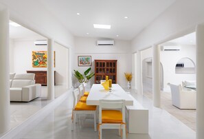 A spacious dining area with a long white table, yellow cushions, and white chairs. There is a wooden cabinet and a painting on the wall. - Beautiful Open Plan Villa On 3,000  of Living Space at No 1 Queens Fort (St James)