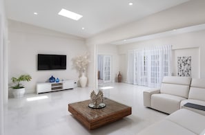A spacious living room with a modern white sofa, a glass coffee table with a decorative centerpiece, a flat-screen TV mounted on the wall, and a white console table with decorative items. - Beautiful Open Plan Villa On 3,000  of Living Space at No 1 Queens Fort (St James)