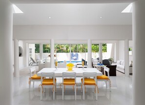 A modern dining area with a white table and chairs, a yellow centerpiece, and a view of a pool and outdoor seating. - Beautiful Open Plan Villa On 3,000  of Living Space at No 1 Queens Fort (St James)