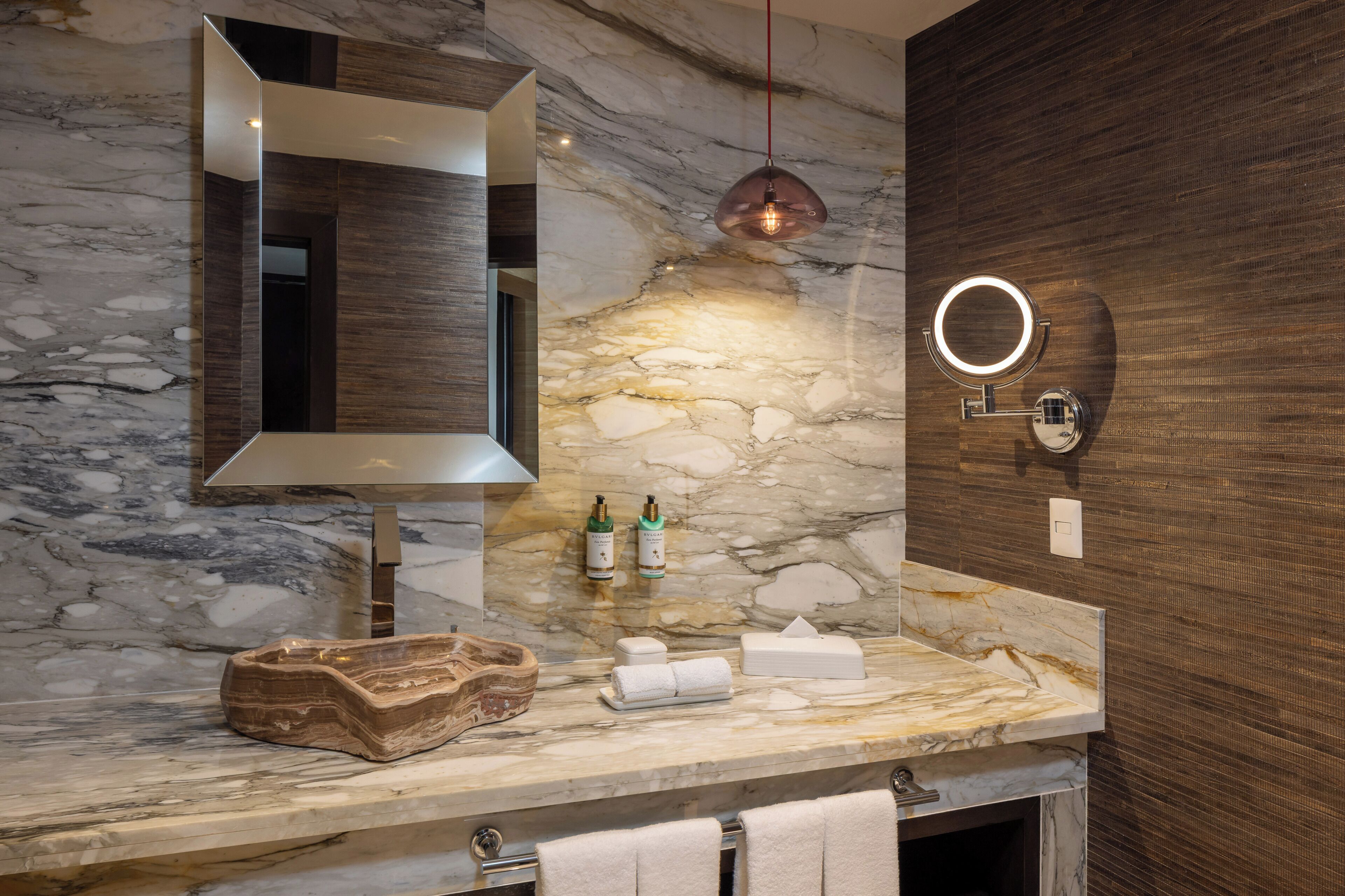 A bathroom with a marble countertop, a large mirror, a pendant light, a round mirror, and toiletries.