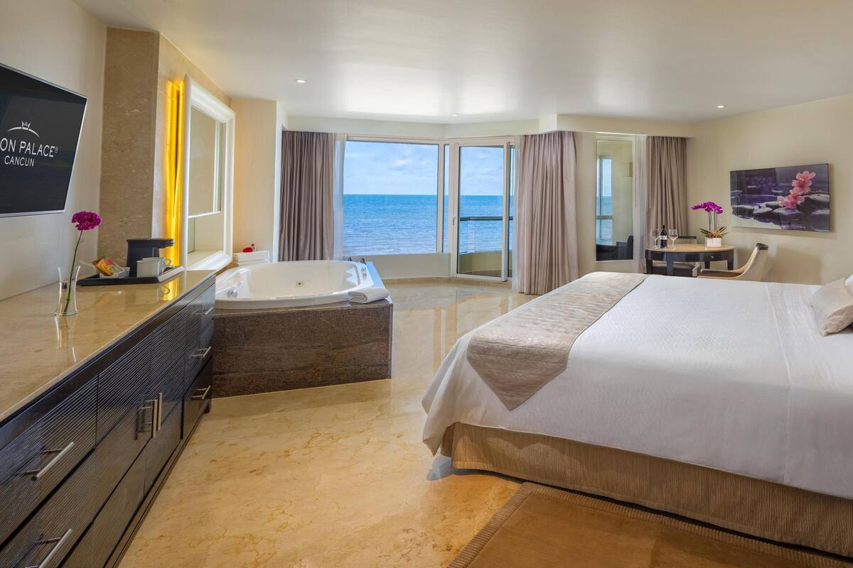 Habitación de hotel con una cama grande, una bañera, tocador con espejo y vista al océano.
