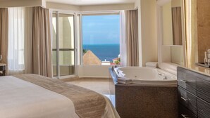 A hotel room with a large bed, a bathtub, a bathroom with a mirror, and a view of the ocean.