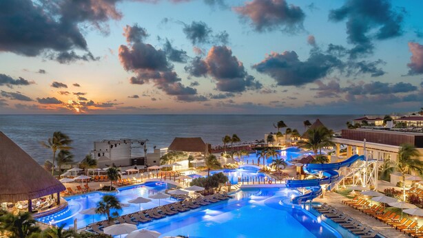 A resort pool area with a water slide, lounge chairs, and a sunset view.