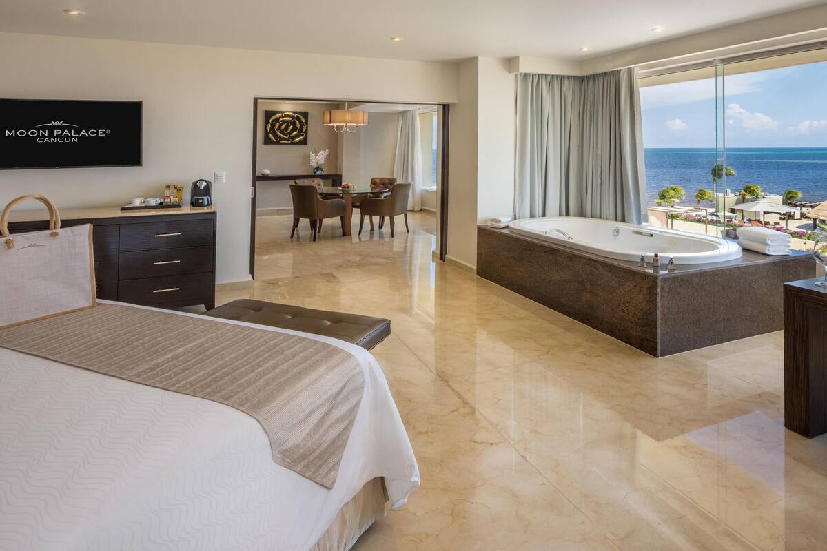 Habitación de hotel moderna con una cama grande, un área de estar con vista al océano, un balcón privado con piscina y un televisor mostrando el nombre del hotel.