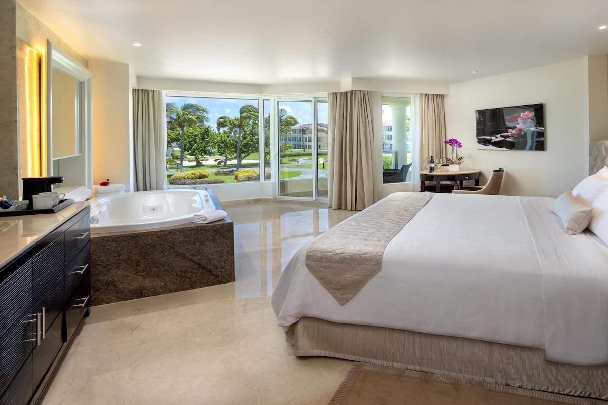 Habitación de hotel con una cama grande, un escritorio, una silla, un televisor y un baño con bañera y un ventanal amplio.
