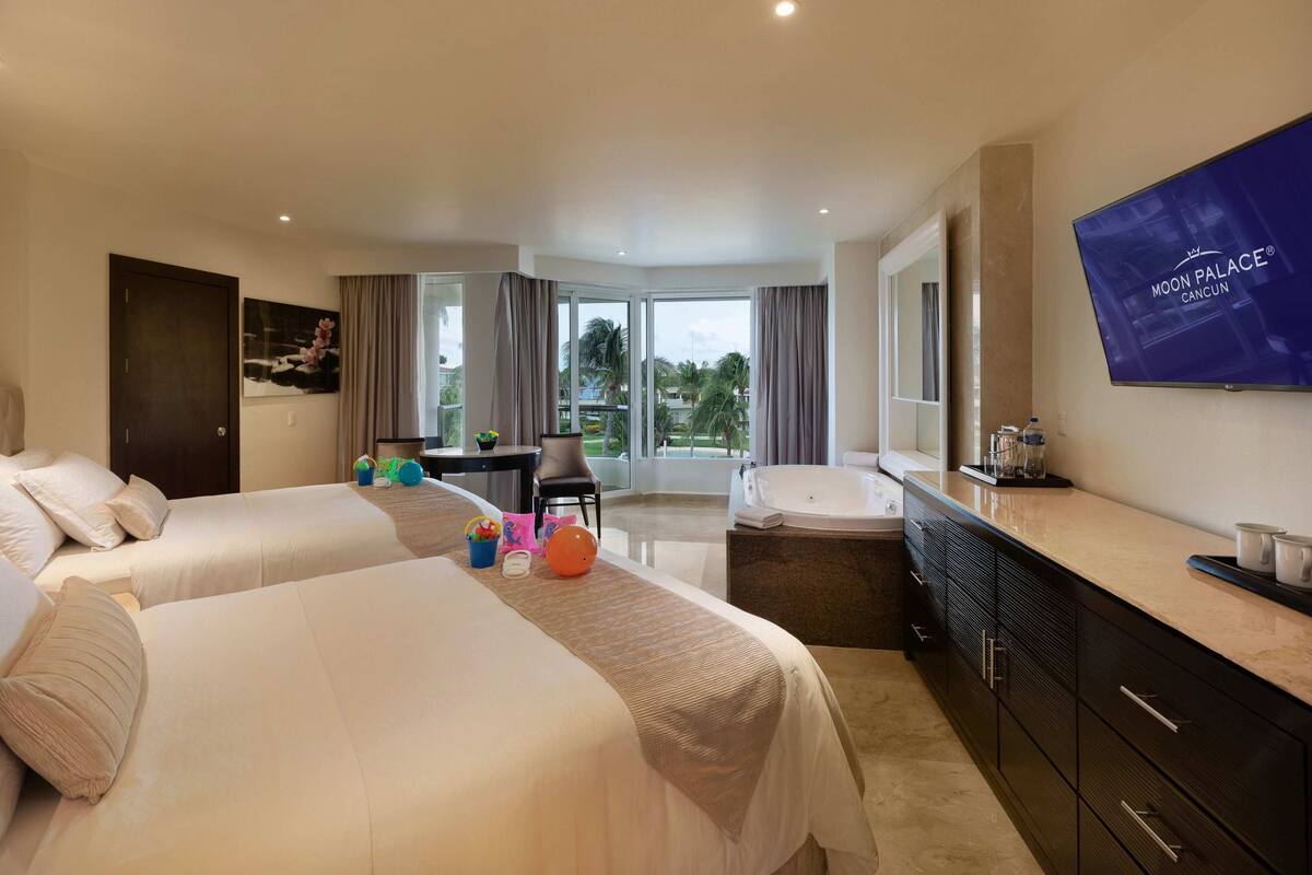 Habitación de hotel con dos camas, un ventanal grande, televisor, baño y balcón.