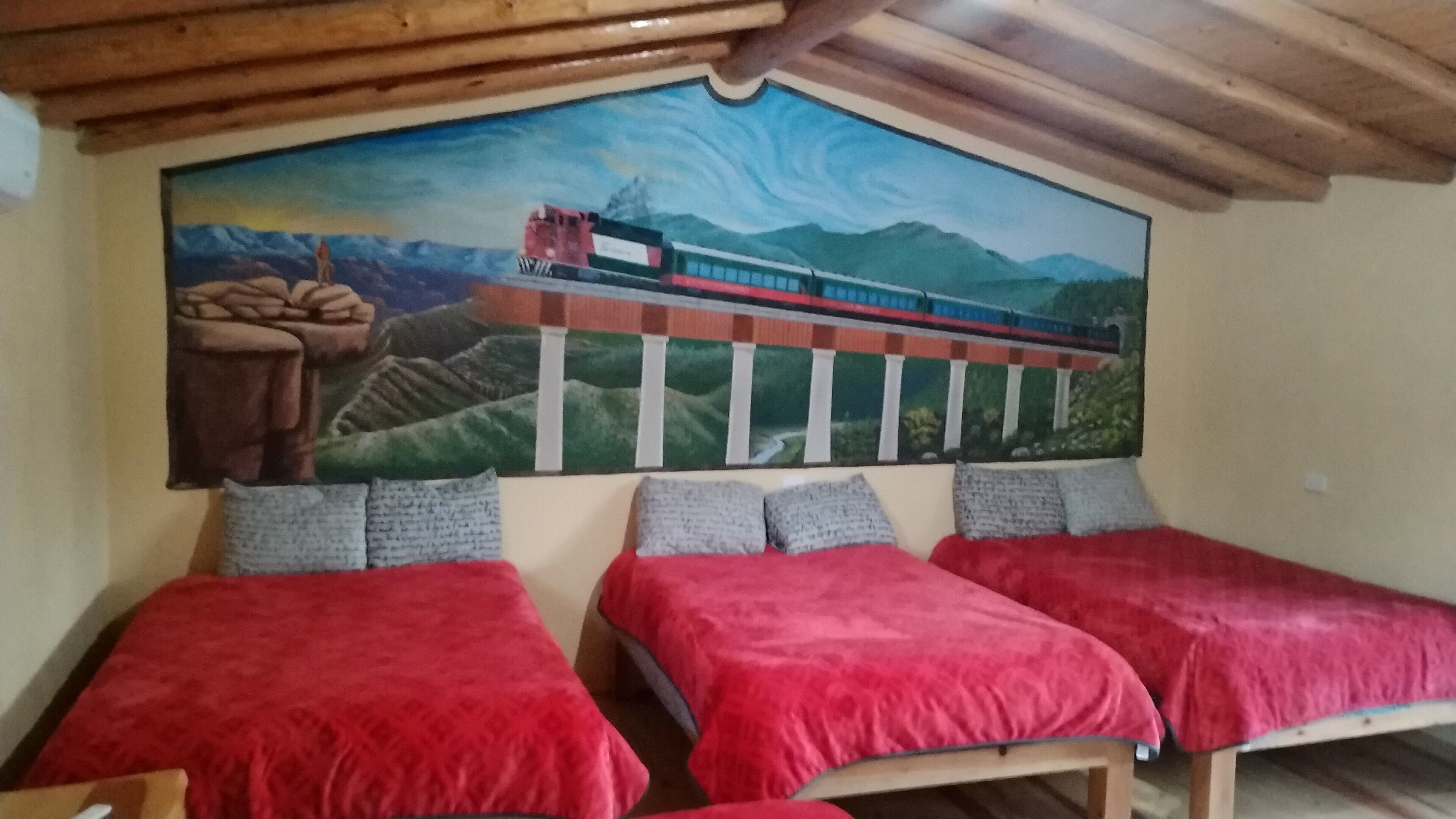 Une pièce avec deux lits recouverts de couvertures rouges, une fresque représentant un train sur un pont et un plafond en bois.