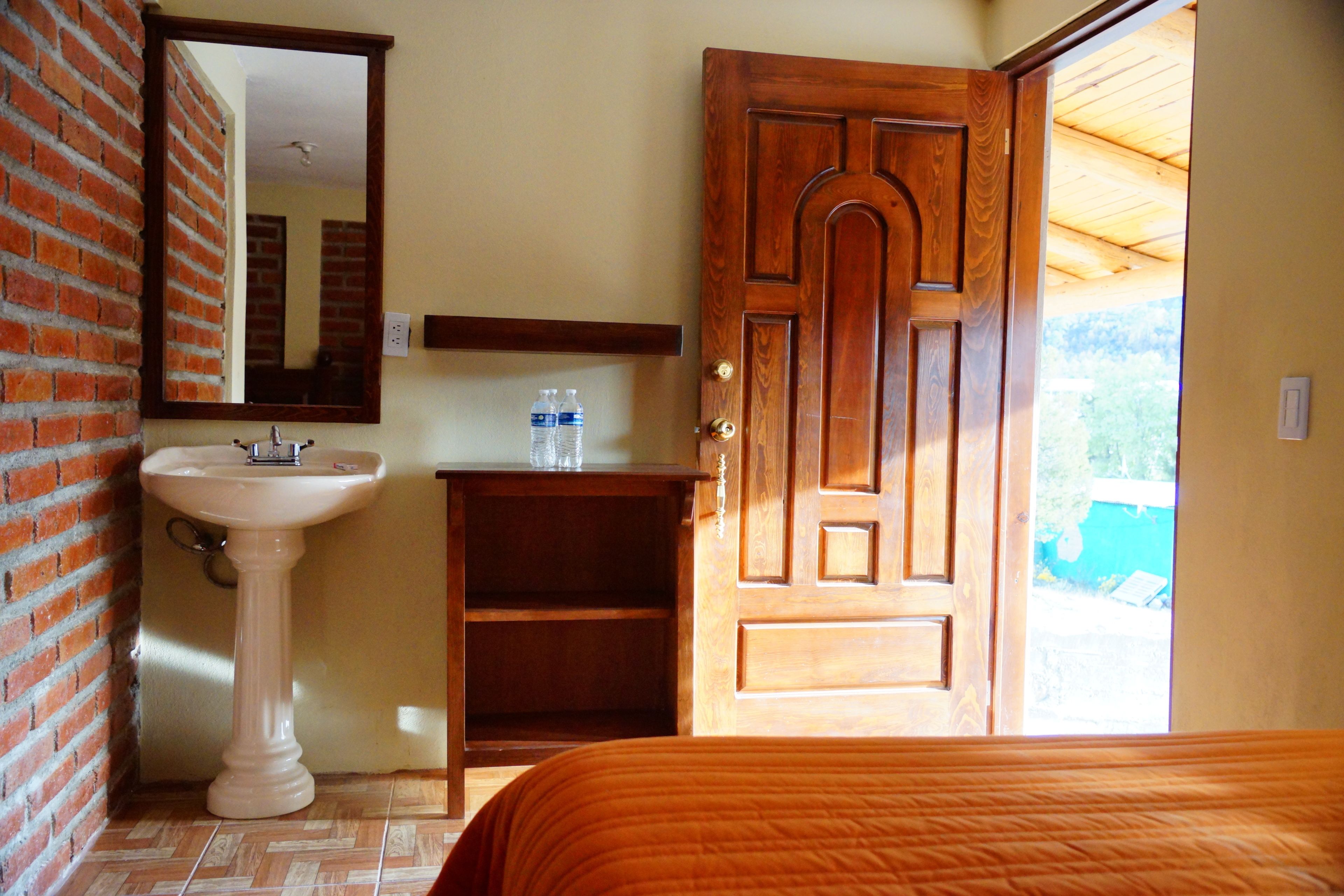 Un baño con lavamanos pedestal, espejo, puerta de madera y cama con colcha naranja.