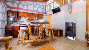 A rustic kitchen with wooden cabinets, a stone wall, a wooden dining table, and a black stove. - Cabañas Las Gemelas Barrancas del Cobre (Urique)