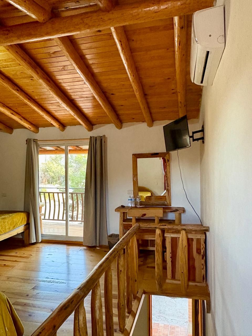 Habitación con piso de madera, balcón, cama, escritorio y televisor montado.
