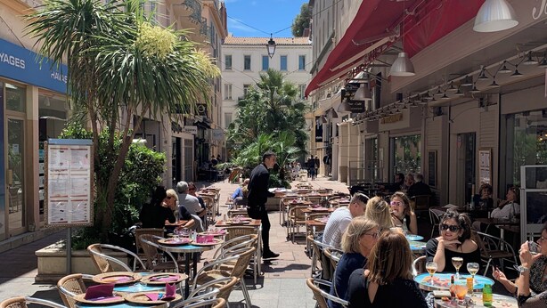 Un café al aire libre con mesas y sillas, rodeado de edificios con balcones y toldos.