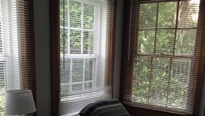 A room with two single beds, a round table with a lamp, and large windows with blinds.