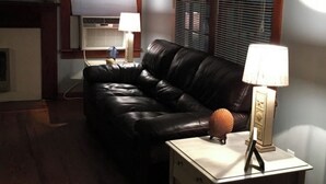 A living room with a black leather sofa, a small white table with a lamp, and two windows with blinds.