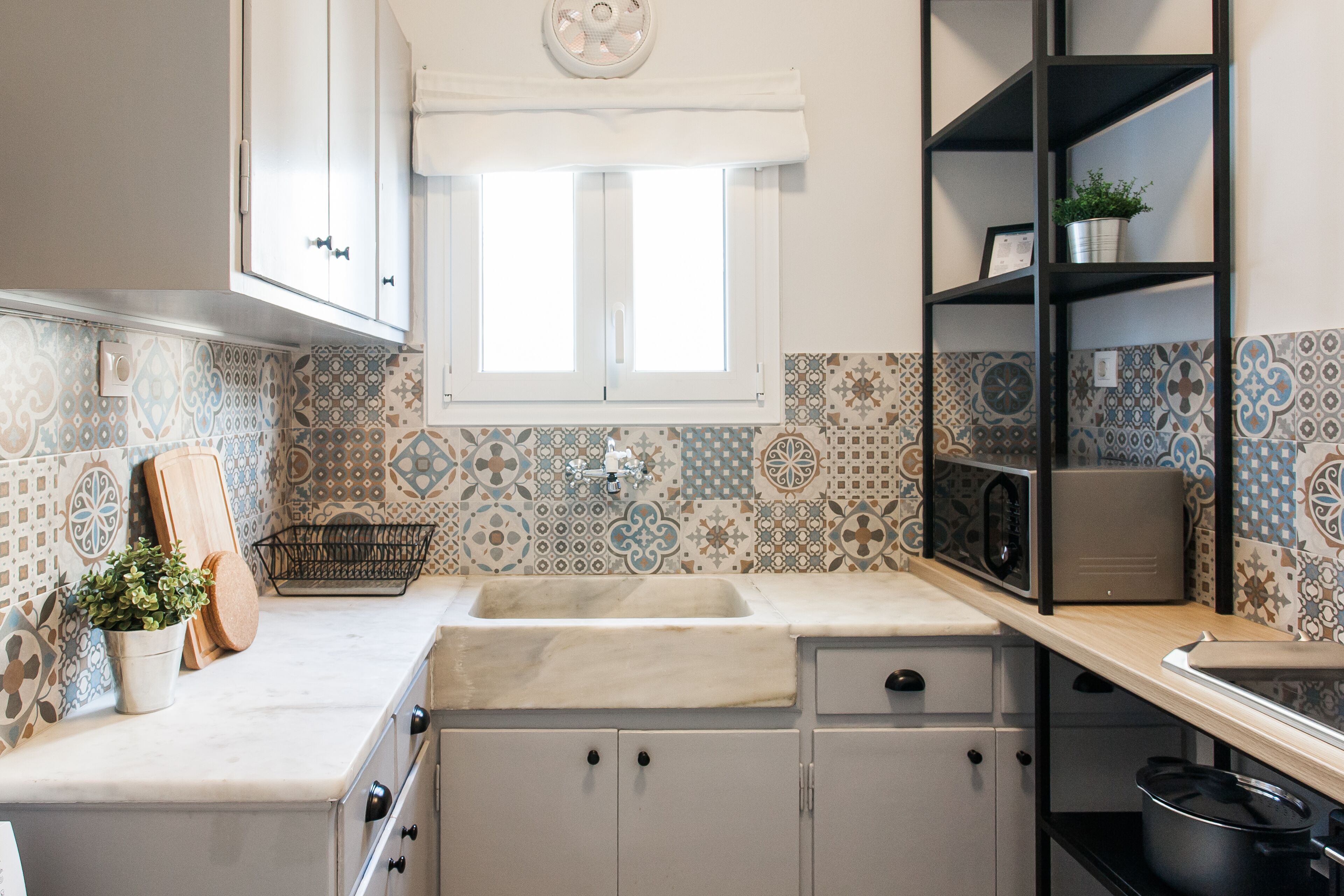 A kitchen with a window, patterned tiles, a sink, cabinets, and a shelf with a plant.