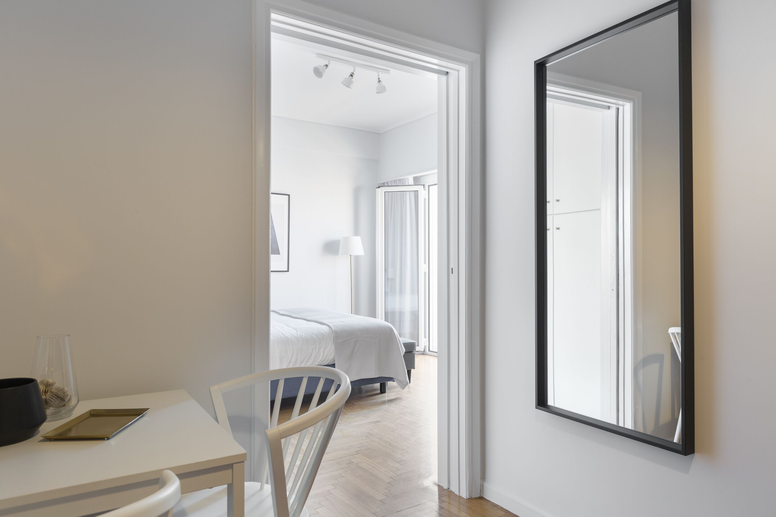 A modern hotel room with a large mirror, a bed with white bedding, a bedside table with a lamp, and a view of the hallway.