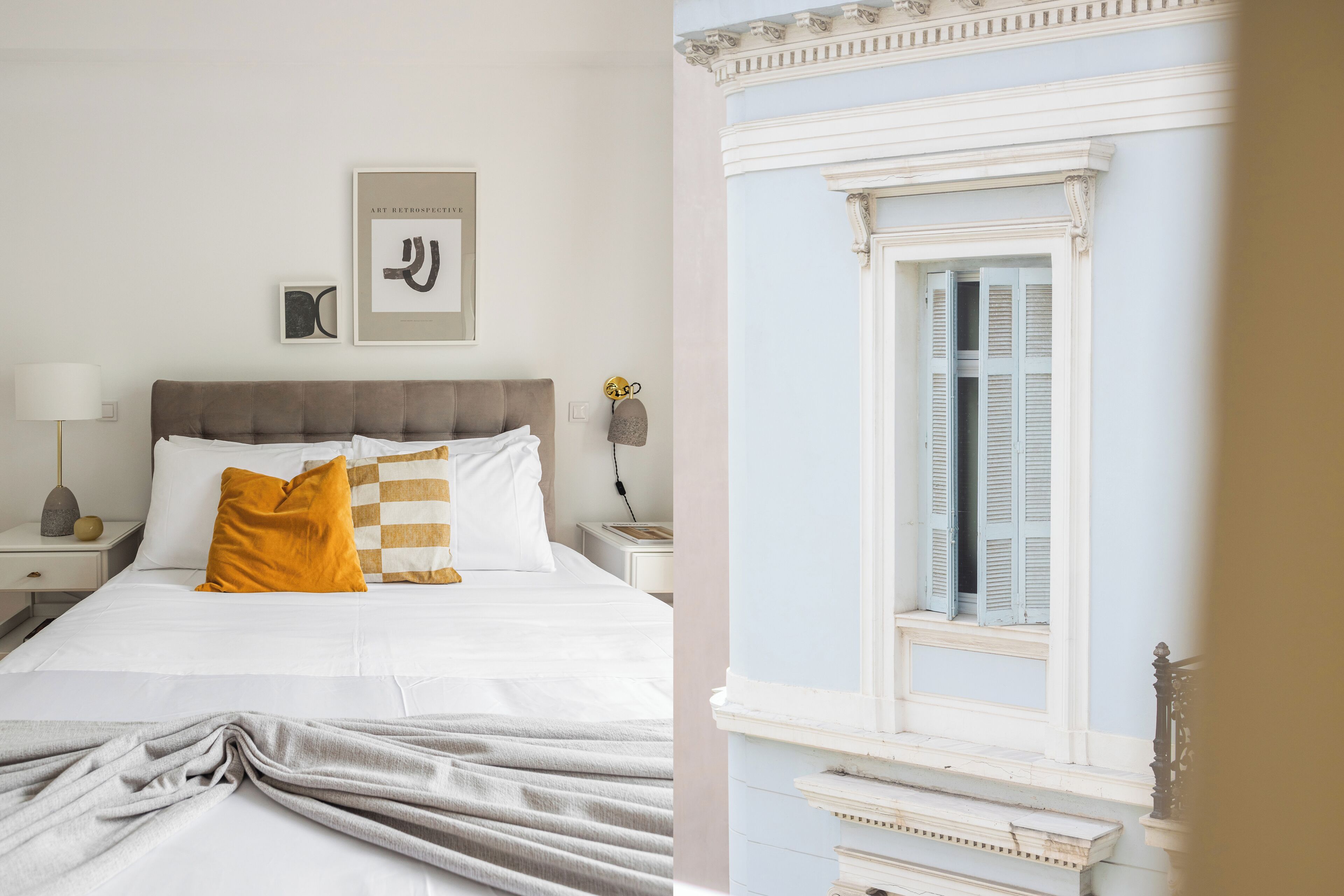 A bedroom with a bed, white bedding, a gray throw blanket, a nightstand with a lamp, and a framed picture on the wall.