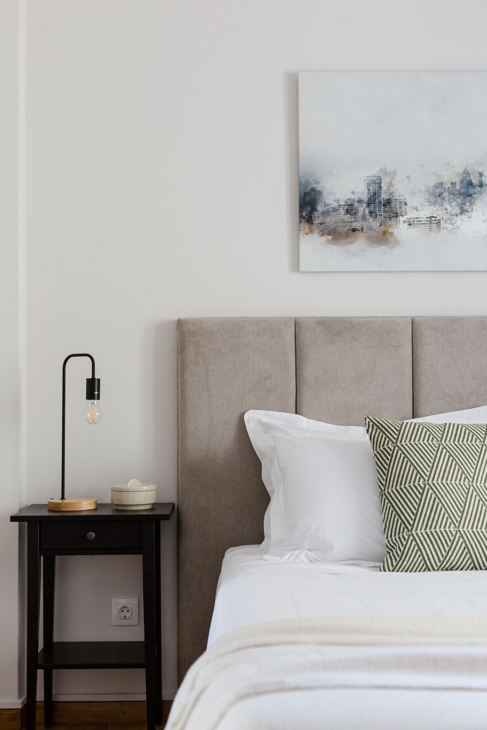 Quarto com cama, mesa de cabeceira, abajur e um quadro na parede.