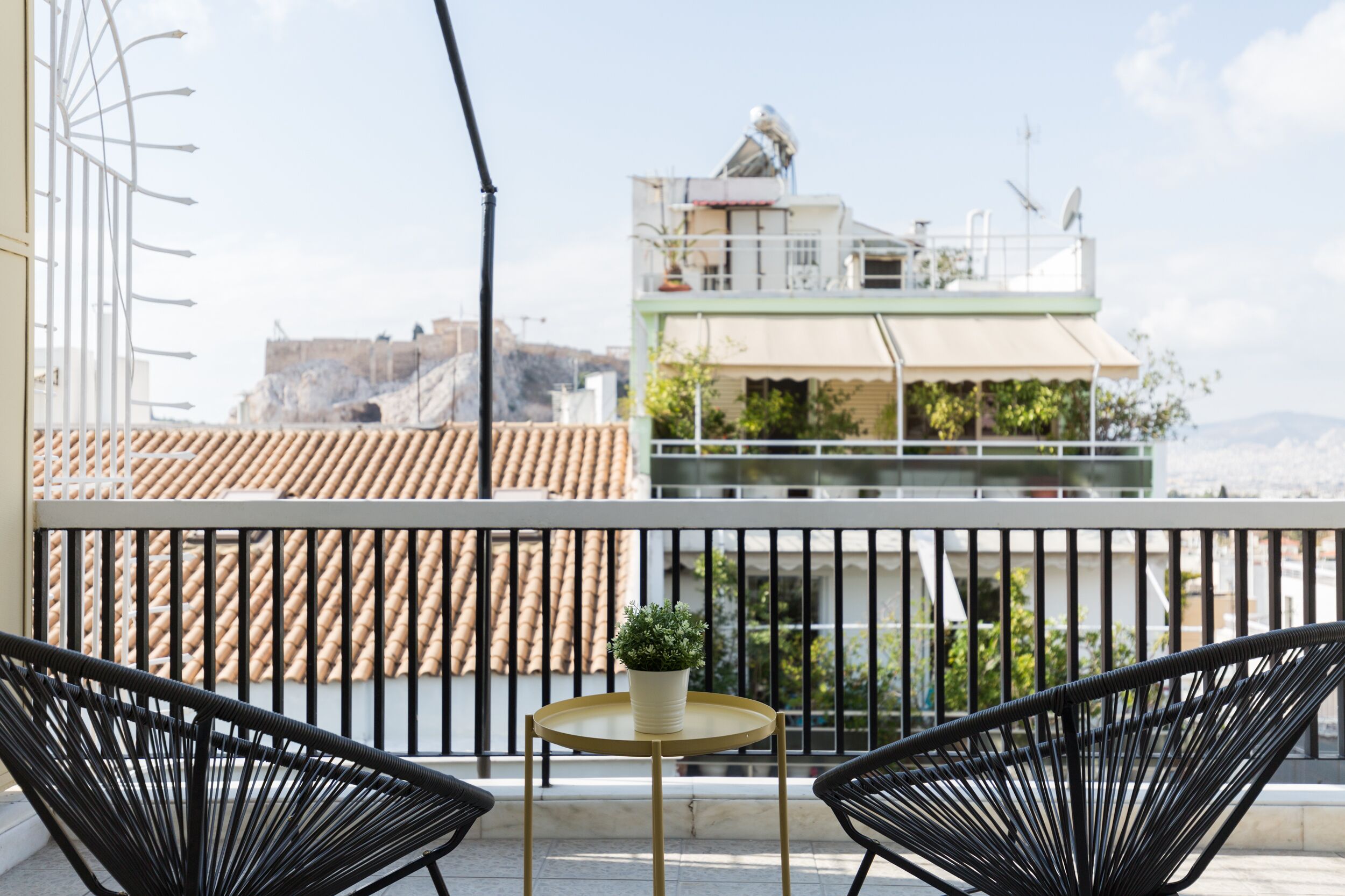A balcony with two black wicker chairs, a small round table with a potted plant, and a view of buildings in the background.