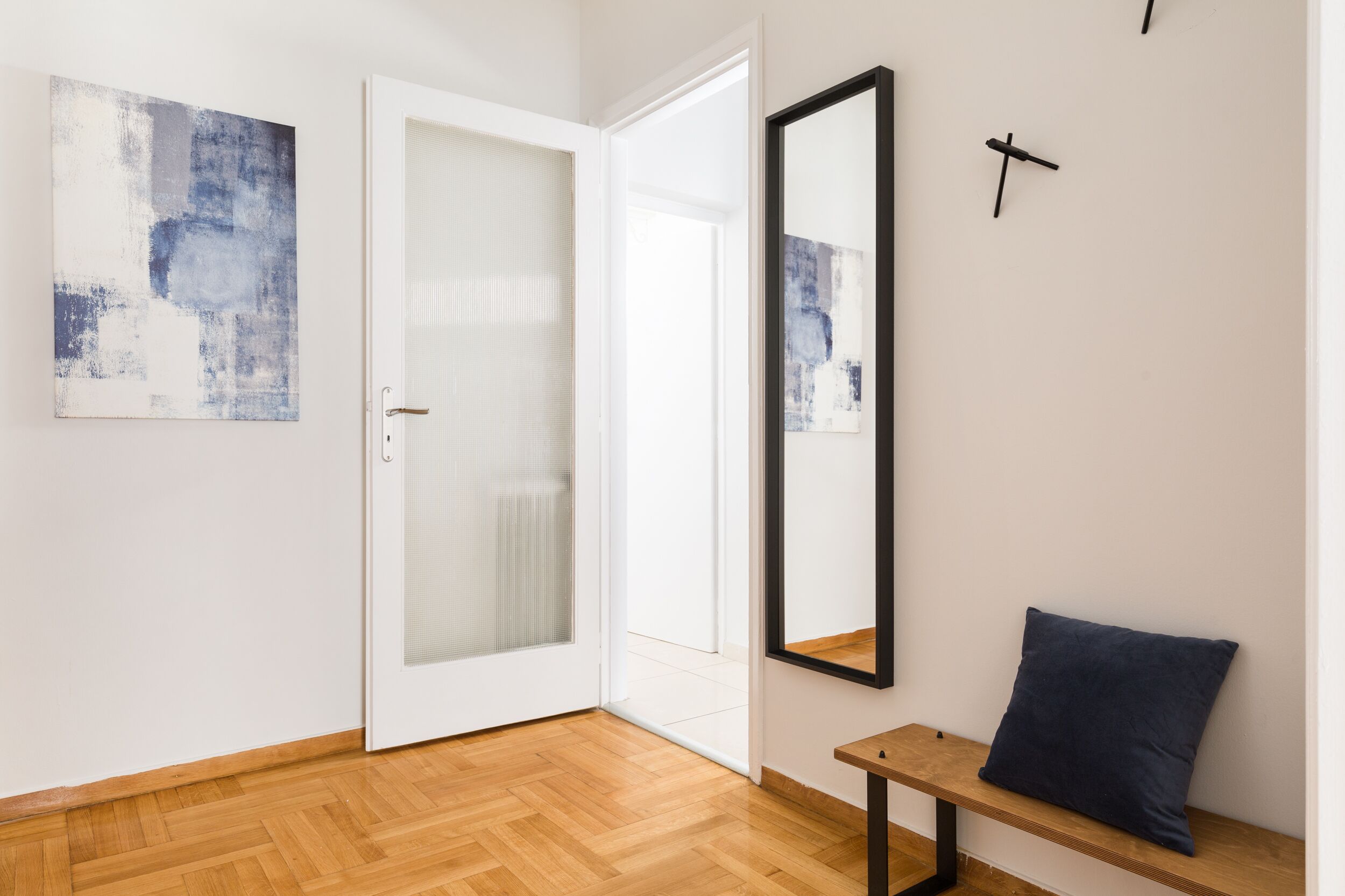 A room with a wooden floor, a bench with a cushion, a door with a frosted glass panel, and two abstract paintings on the wall.
