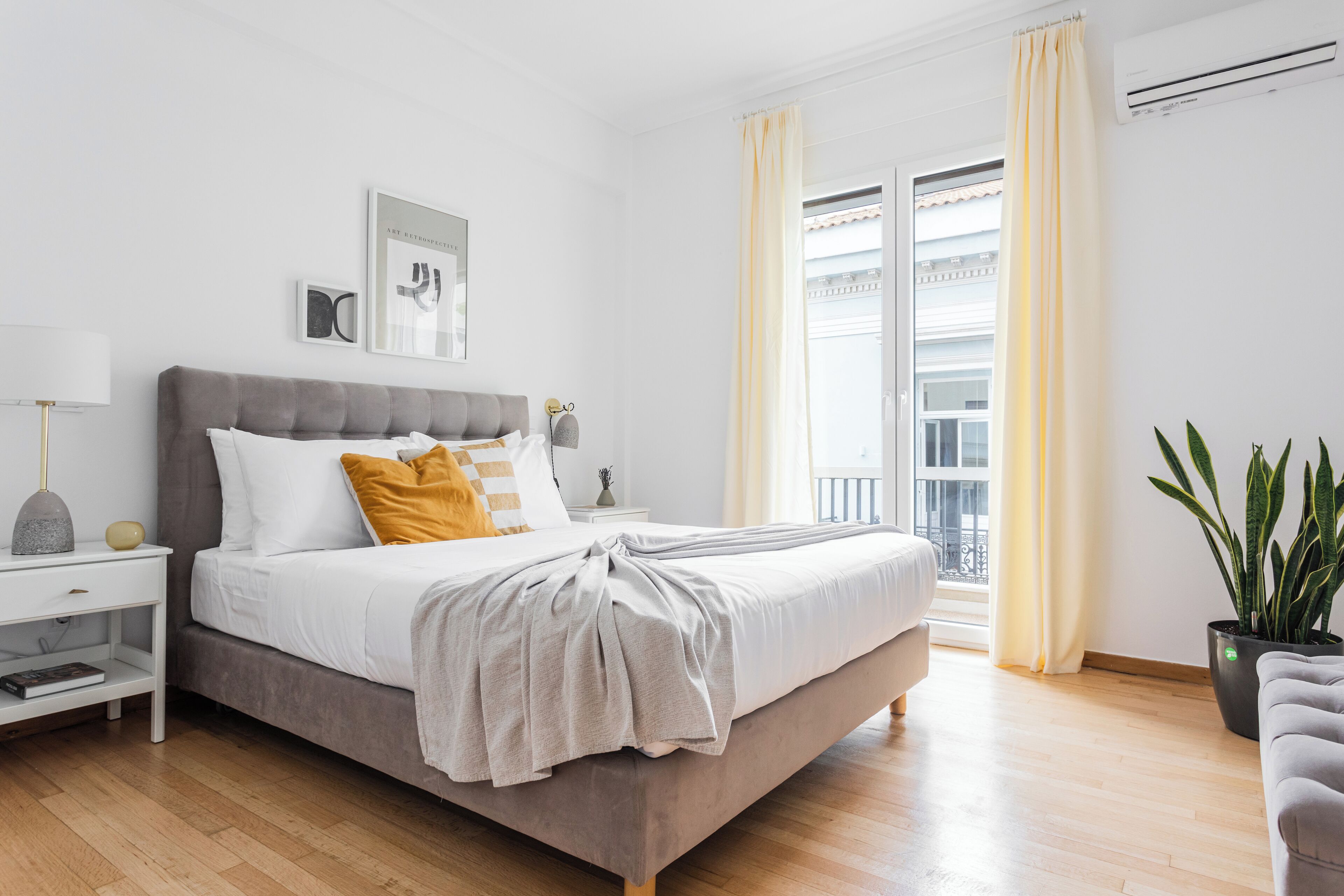 A modern bedroom with a large bed, a bedside table with a lamp, a potted plant, and a view of the outside balcony.