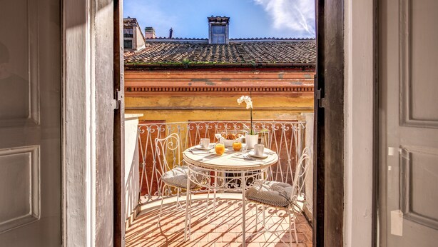 An open balcony with a table set for two, a potted plant, and a view of a building with a tiled roof. - Maison Babuino with a panoramic terrace (Roma)