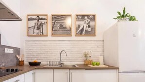 Una cocina moderna con armarios blancos, un lavamanos de acero inoxidable y un refrigerador blanco. Hay tres cuadros enmarcados en la pared sobre el lavamanos.
