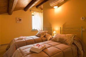 Un dormitorio con dos camas, cada una con edredón y almohadas, una mesita de noche con lámpara y una ventana con cortinas.