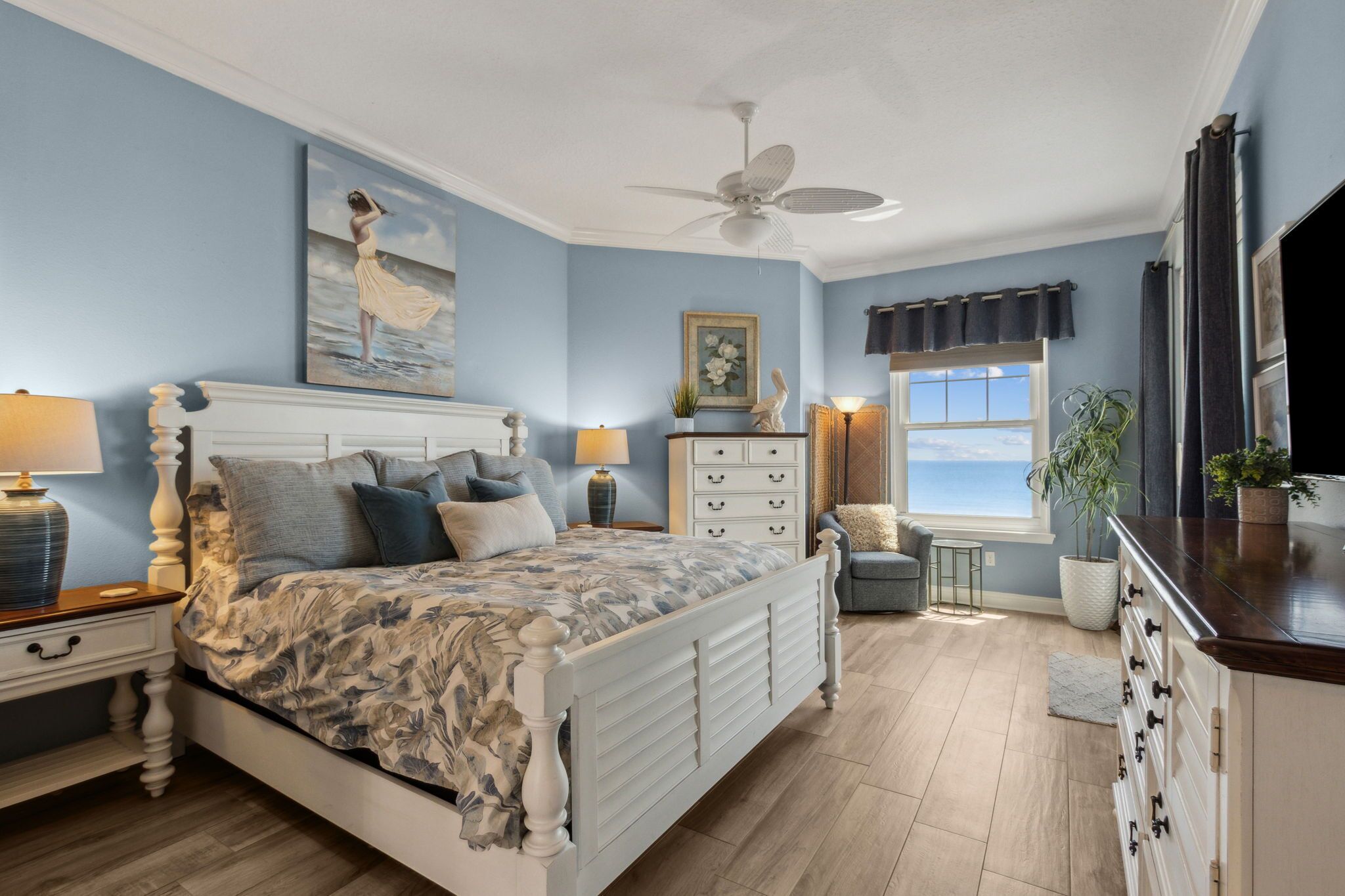 A bedroom with a bed, nightstands, a dresser, a ceiling fan, a window with curtains, and a painting on the wall.
