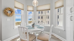 A dining area with a round table, white chairs, and a view of the beach through large windows with blinds.