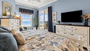 A bedroom with a large bed, a ceiling fan, a dresser, a TV, and a view of the outdoors.