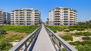 A wooden boardwalk leading to a beachfront hotel with multiple balconies and a clear blue sky.