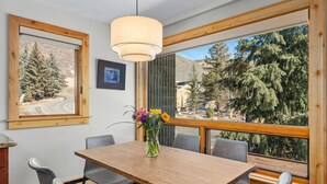 A dining area with a wooden table and grey chairs, a vase with flowers, and a large window with a view of trees and mountains. - Vail 21 303 - 2 Bedroom Platinum (Vail)