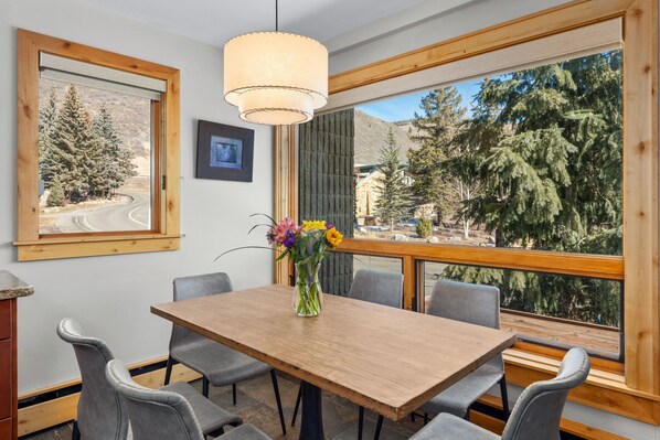 A dining area with a wooden table and grey chairs, a vase with flowers, and a large window with a view of trees and mountains. - Vail 21 303 - 2 Bedroom Platinum (Vail)