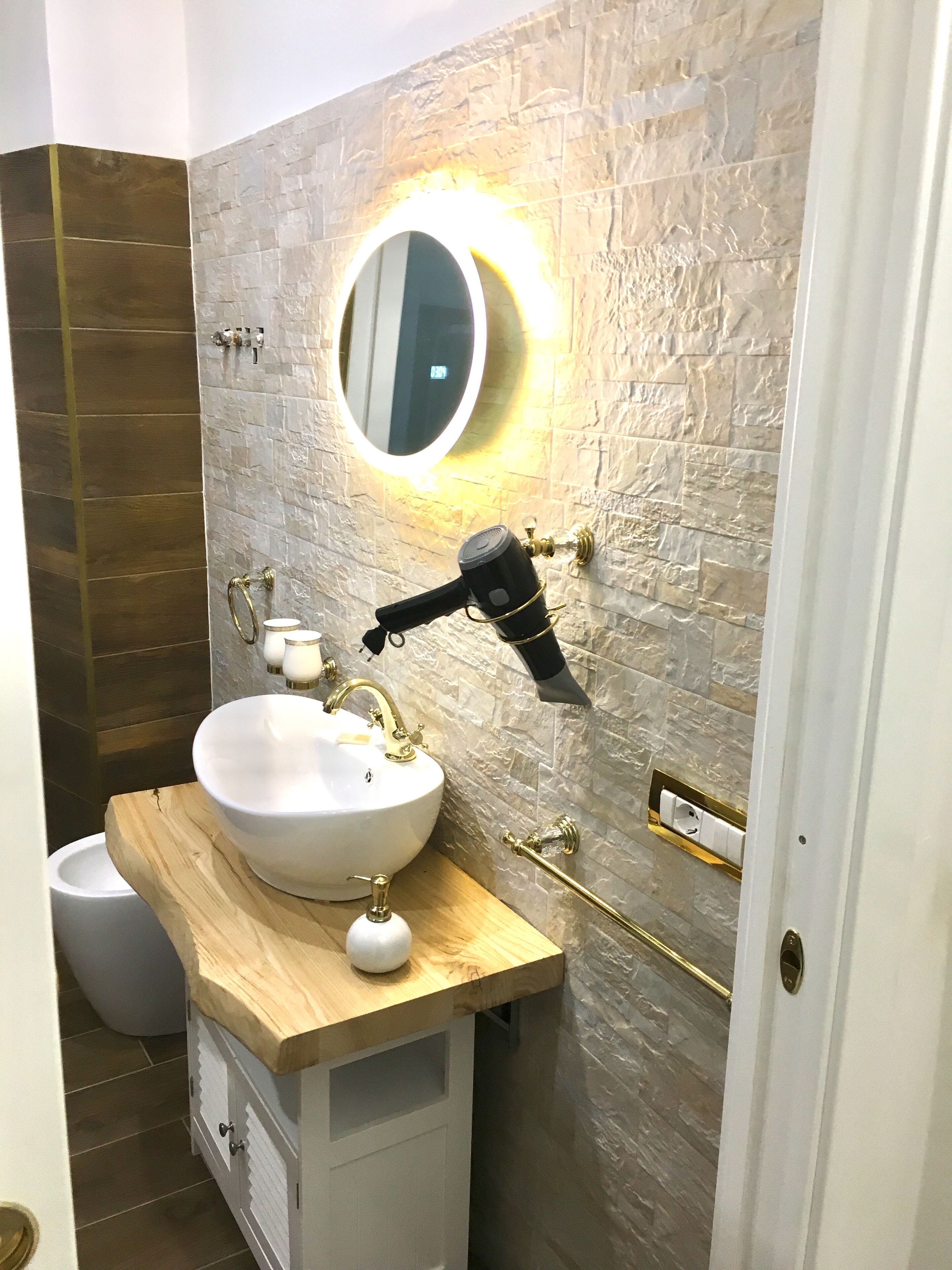 Um banheiro com pia branca, detalhes dourados, um espelho redondo com iluminação e um secador de cabelo fixado na parede.