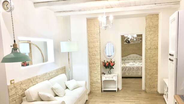 Een kamer met een witte bank, een klein wit tafeltje met een bloevas, een spiegel en een bed op de achtergrond.