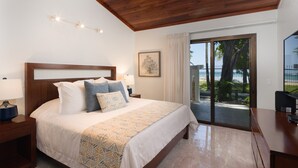 Ein Schlafzimmer mit einem großen Bett, einem Holzkopfende, Nachttischlampen, einem gerahmten Kunstwerk und Blick auf den Strand durch eine Schiebetür.