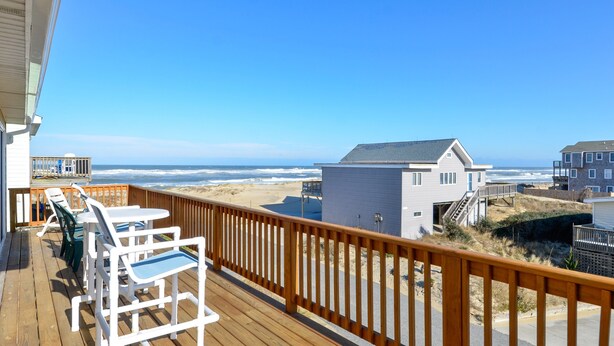 Eine Holzterrasse mit weißen Stühlen und Blick auf Strand und Meer.