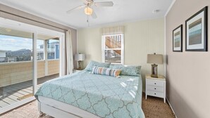 Ein Schlafzimmer mit Bett, Nachttisch, Lampe, Deckenventilator, Fenster mit Jalousien sowie Blick auf einen Balkon und Gebäude.