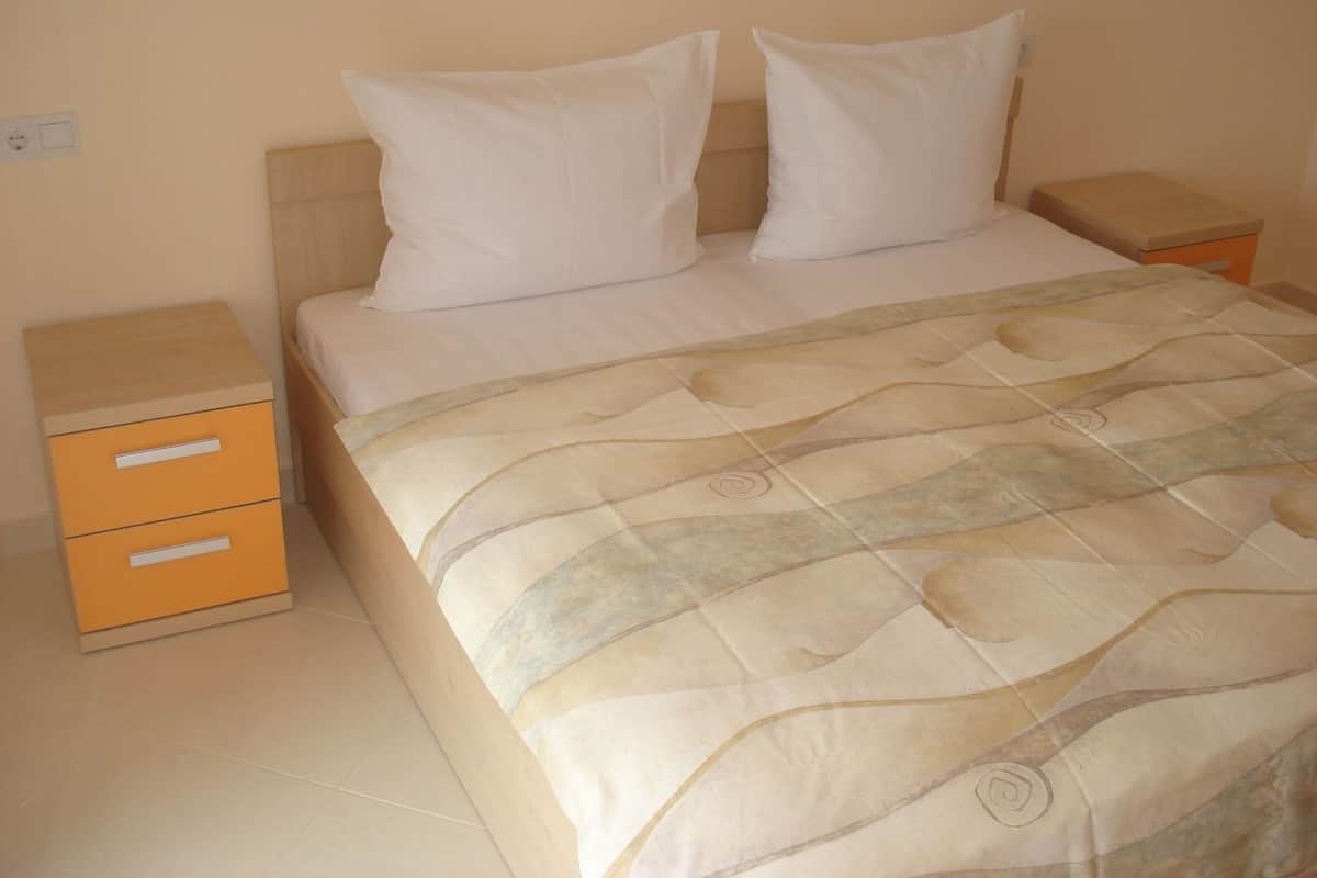 A bed with a patterned bedspread, two white pillows, and a wooden headboard. There are two nightstands with drawers, one of which has a light switch.