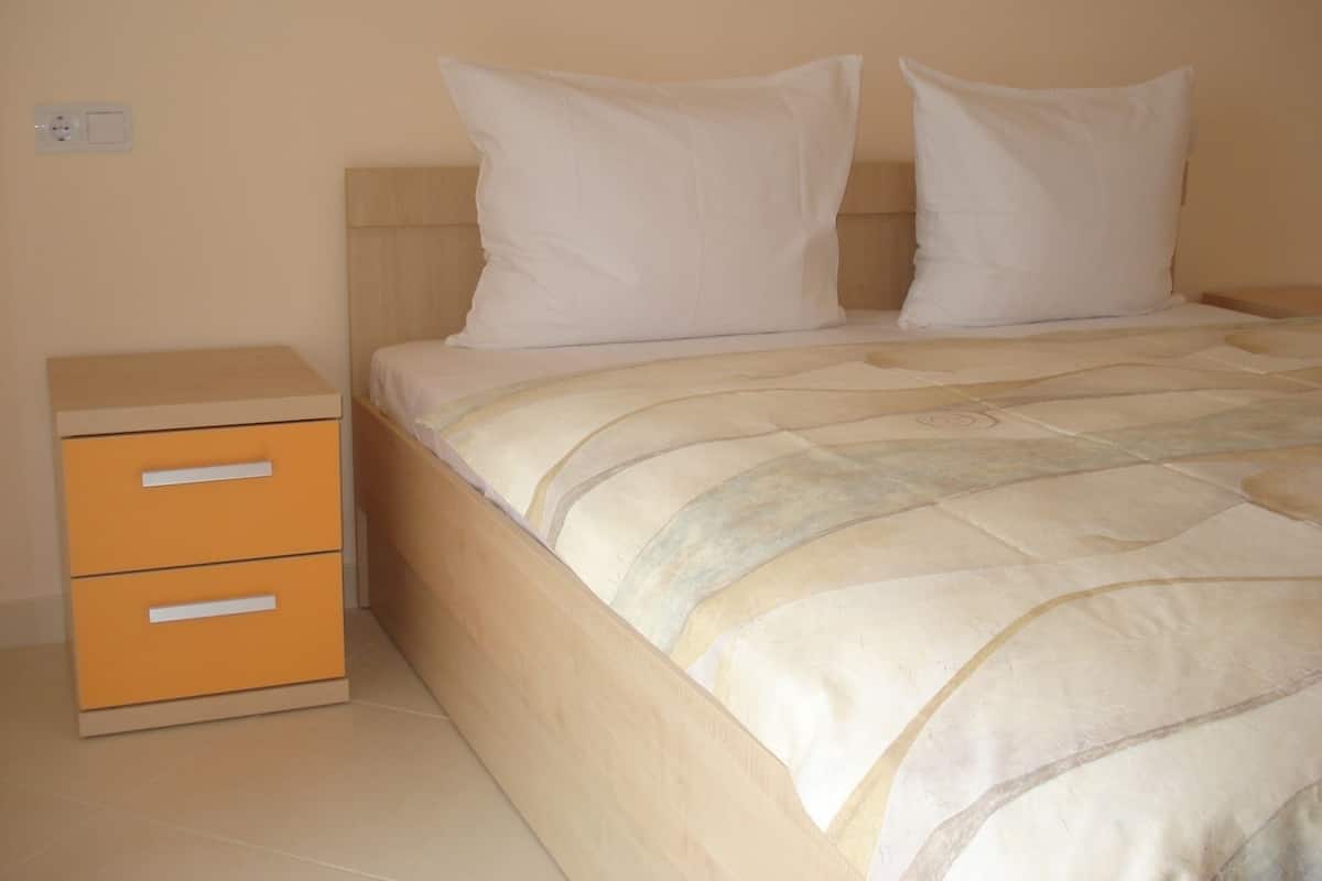 A bedroom with a bed, two pillows, a nightstand with drawers, and a wall socket.