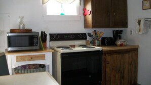 A compact kitchen with a microwave, stove, and oven. There is a window with a curtain, wooden cabinets, and a small countertop with various items.