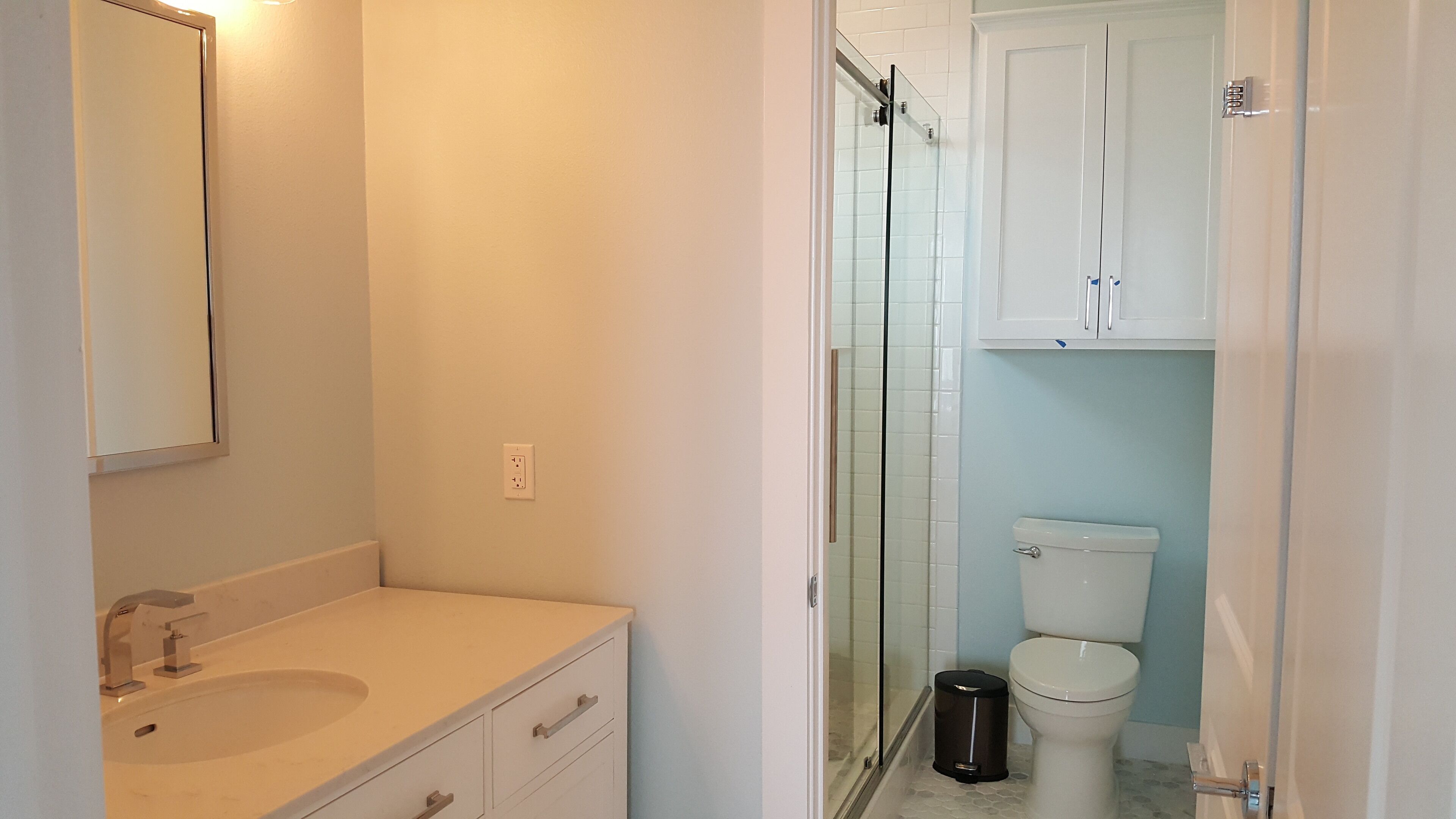 Un baño con lavamanos blanco, espejo, y armarios, así como una cabina de ducha de vidrio y un inodoro.