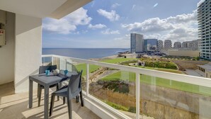 A balcony with a table and chairs, overlooking a cityscape and the sea.