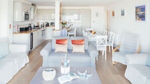 Una sala de estar moderna con un sofá gris, sillones blancos y una mesa de centro. Se ve una cocina al fondo con armarios blancos y un comedor con una mesa y sillas blancas.