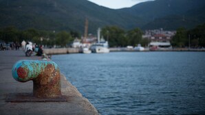 Escena costera con una estructura metálica oxidada, agua tranquila y montañas distantes.