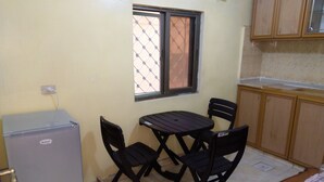 A compact kitchen with a refrigerator, a dining table with four chairs, a window with a metal screen, and wooden cabinets.