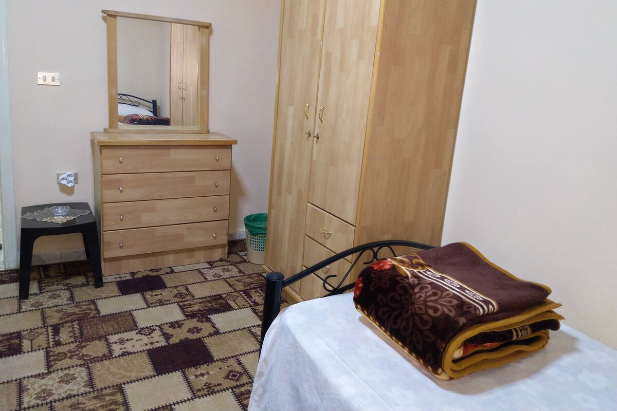 A bedroom with a wooden wardrobe, a small dresser, a mirror, a patterned rug, and a bed with a blanket.